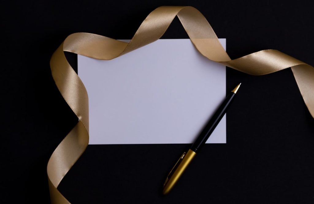 A gold pen, a white piece of paper and a gold ribbon lie on a white background. Top view. Flatley. Layout. Postcard. Letter. Advertising. Place for text. Holiday. The invitation. Wedding